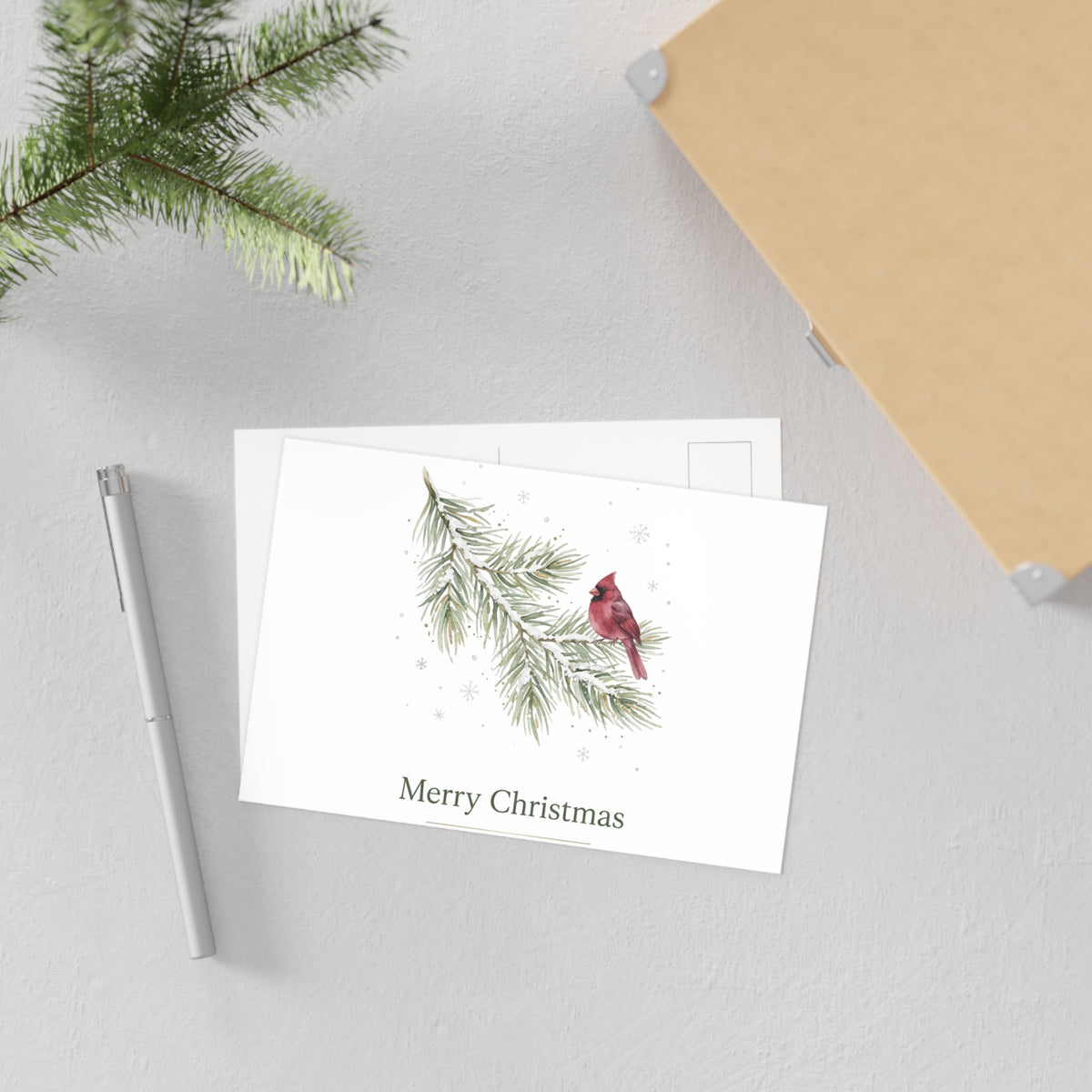 A white Christmas greeting card with a watercolor illustration of a red cardinal on a pine branch and the text 'Merry Christmas,' styled on a light gray surface with a pen, evergreen branch, and notebook corner visible.